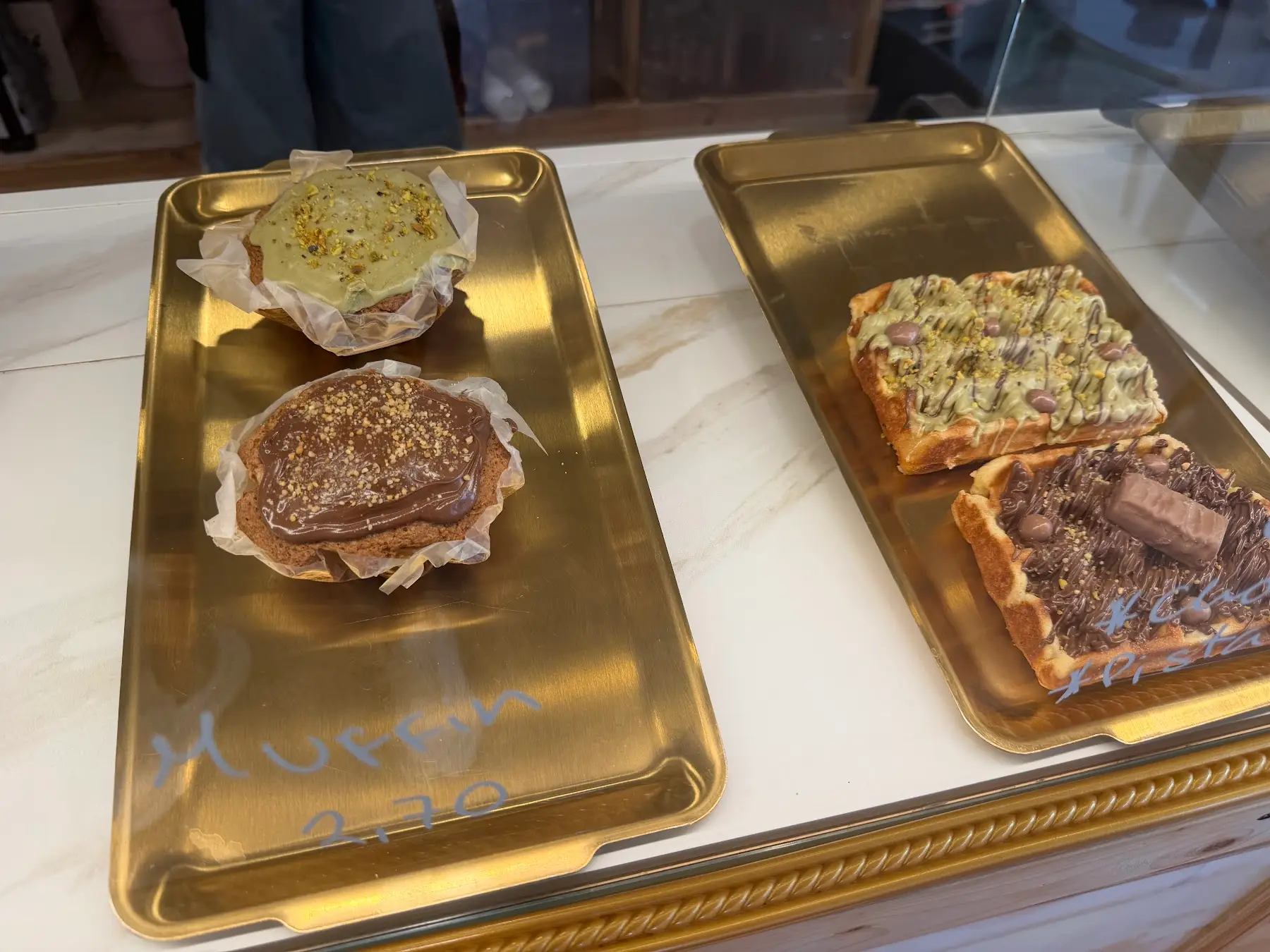 Bandejas doradas en la vitrina con muffins de pistacho y chocolate y gofres con toppings