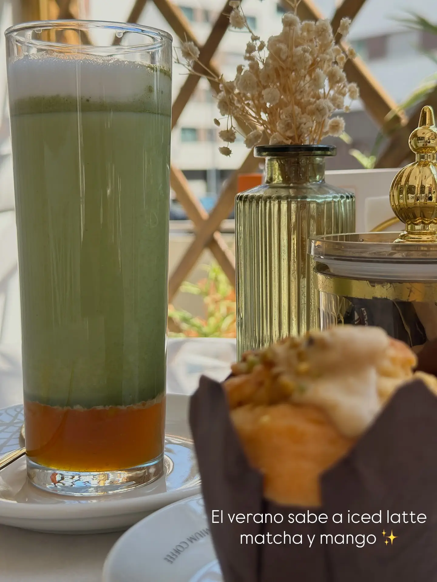 Iced latte de matcha y mango en vaso alto, con el bote dorado y un jarrón de flores secas