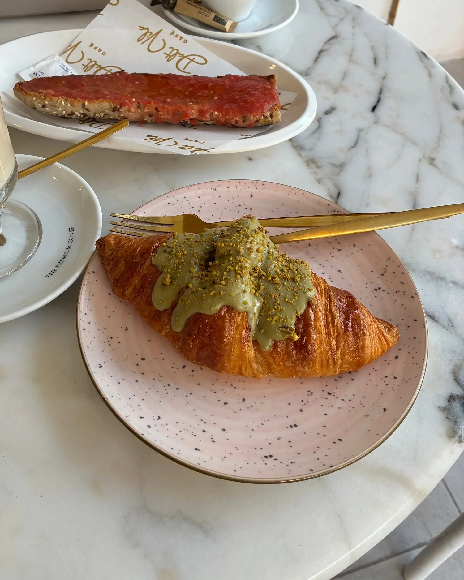 Croissant de pistacho con crema verde sobre un plato rosa moteado, con tenedor dorado, en una mesa de mármol